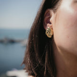 Обеци Спирала с 18К Златно Покритие на жена с къса коса, носеща златисти обеци на фона на море.