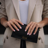 Жена с изискано облекло държи черен портфейл, носейки златисти пръстени и нежно маникюр.