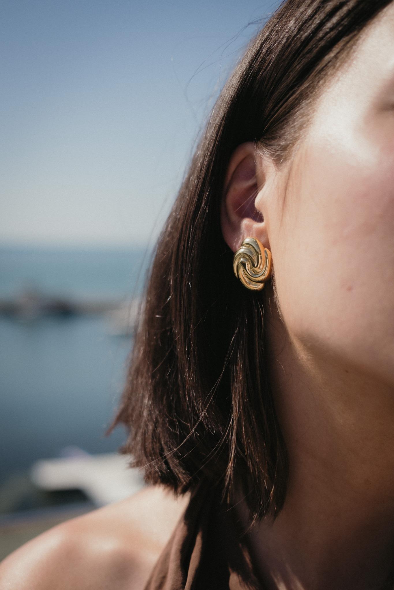 Обеци Спирала с 18К Златно Покритие на жена с къса коса, носеща златисти обеци на фона на море.