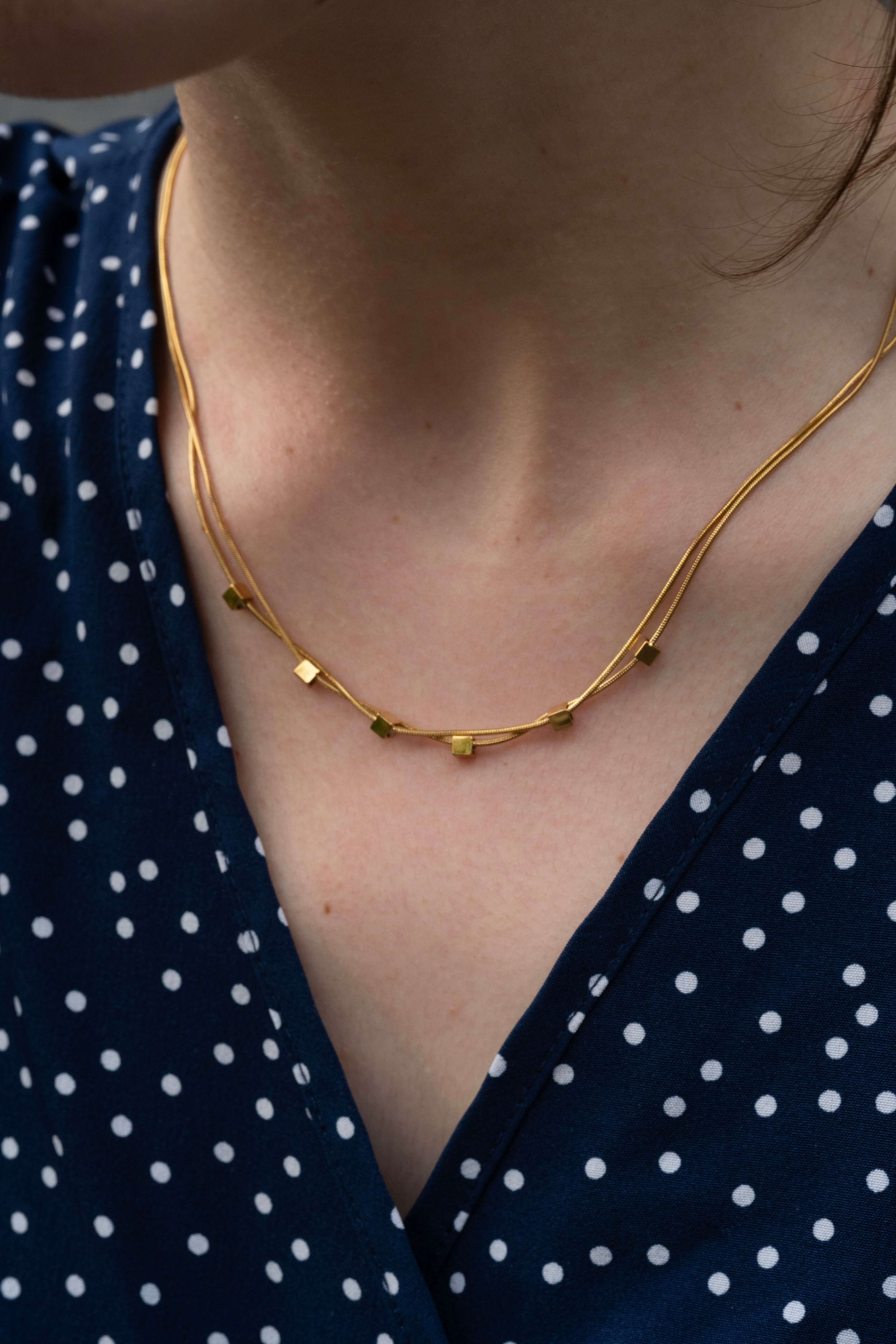 Двойно колие от медицинска стомана с 16к златно покритие и кубчета, носено върху синя риза с бели точки.