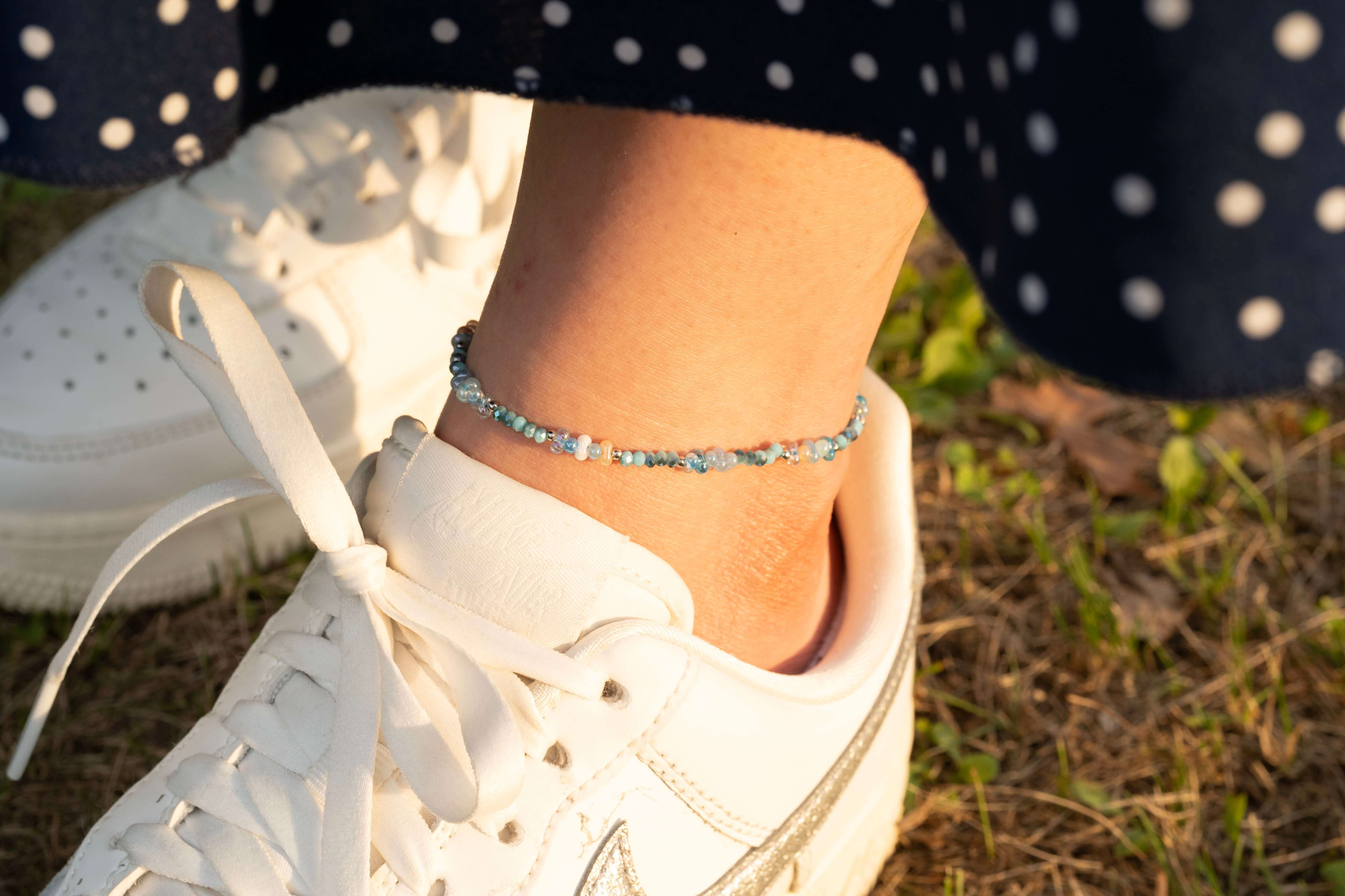 Гривна за глезен с цветни мъниста, съчетана с бяла спортна обувка на фона на тревиста настилка.