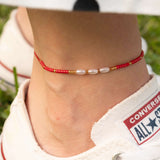 Бохо гривна за глезен от червен конец с перлички, носена с удобни маратонки на зелен фон.