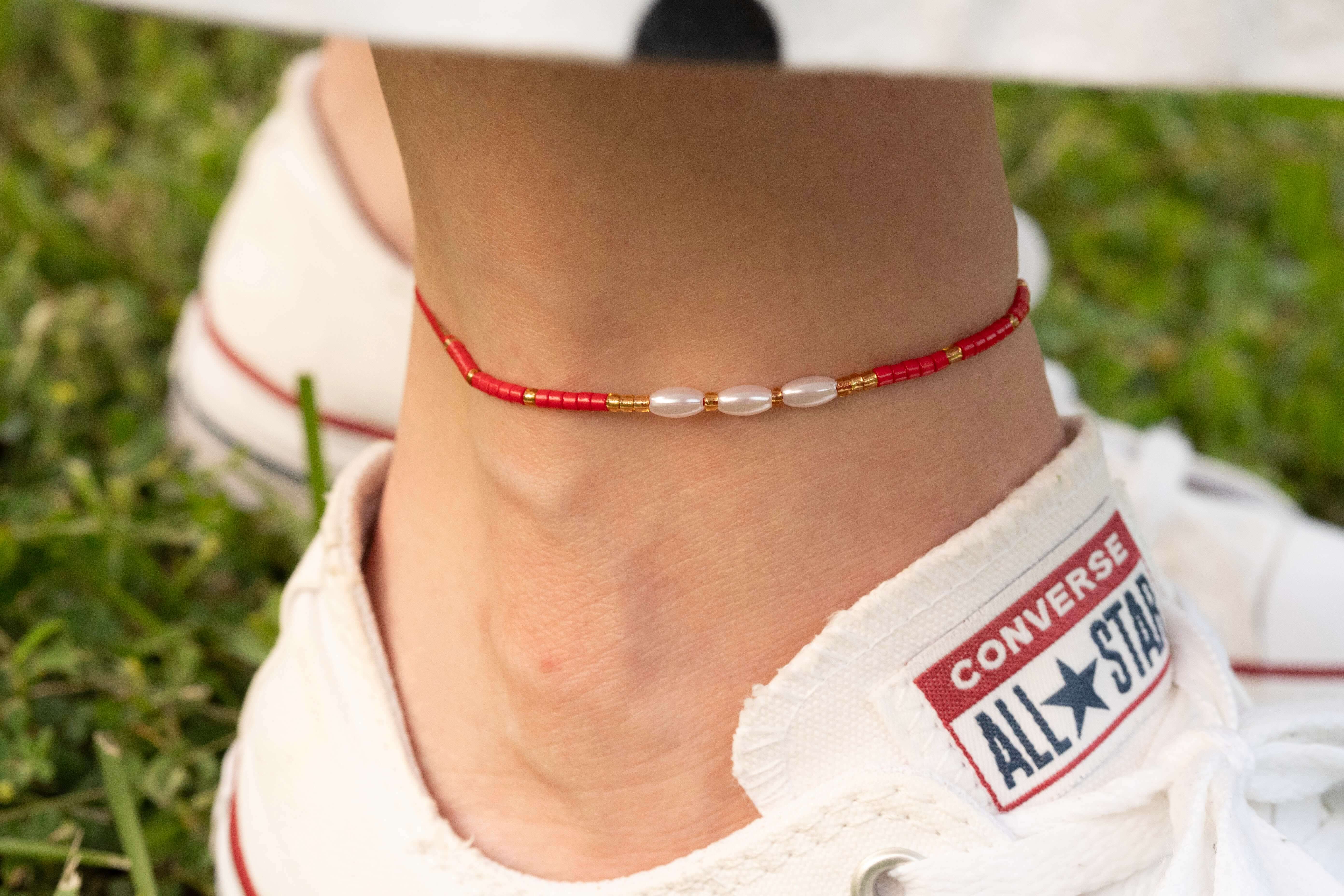 Бохо гривна за глезен от червен конец с перлички, носена с удобни маратонки на зелен фон.