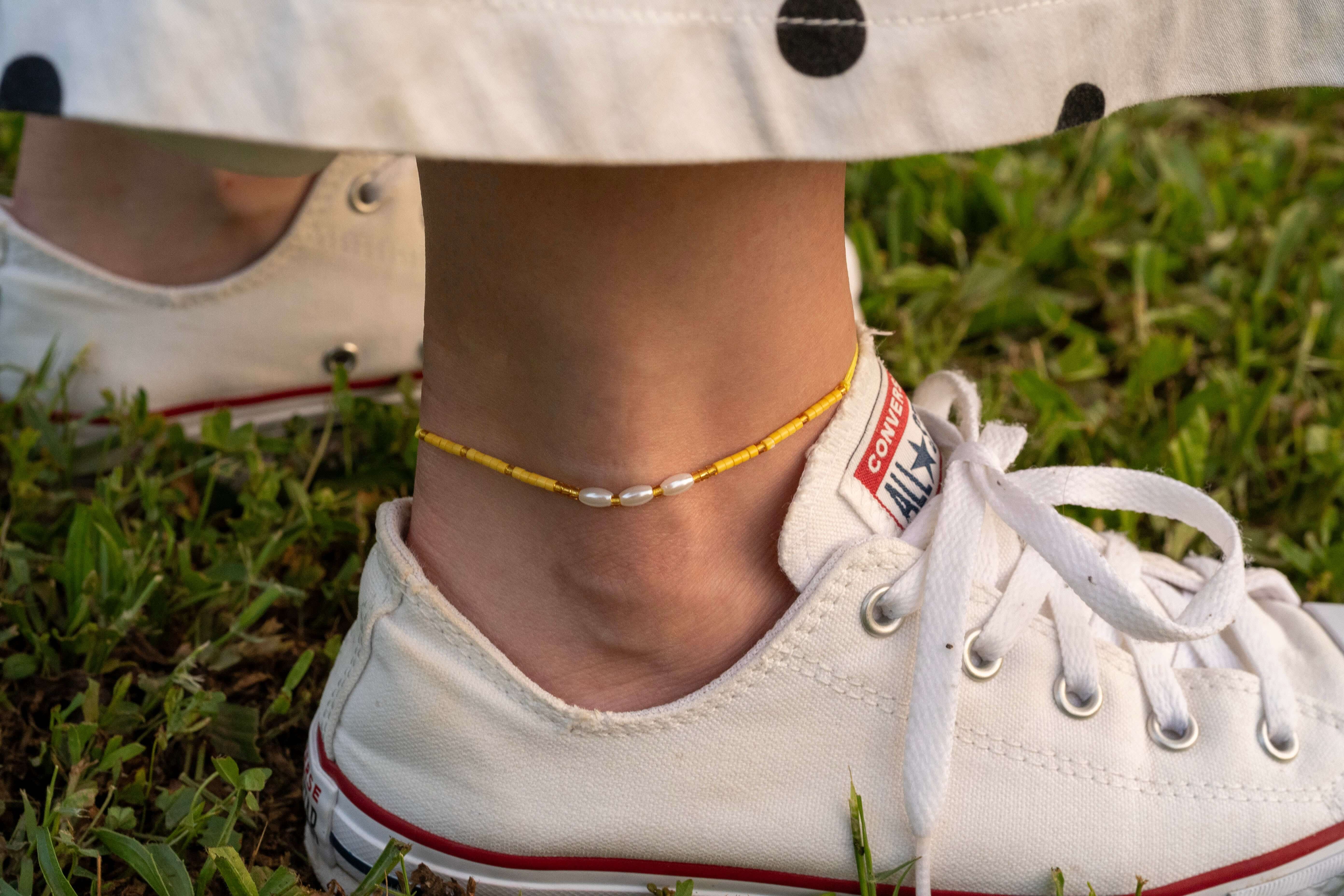 Бохо гривна за глезен с жълти мъниста и перли, носена с бели кеди на зелена трева.