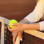 Жена държи тенис топка в ръка, докато се опира на тенис мрежа.
