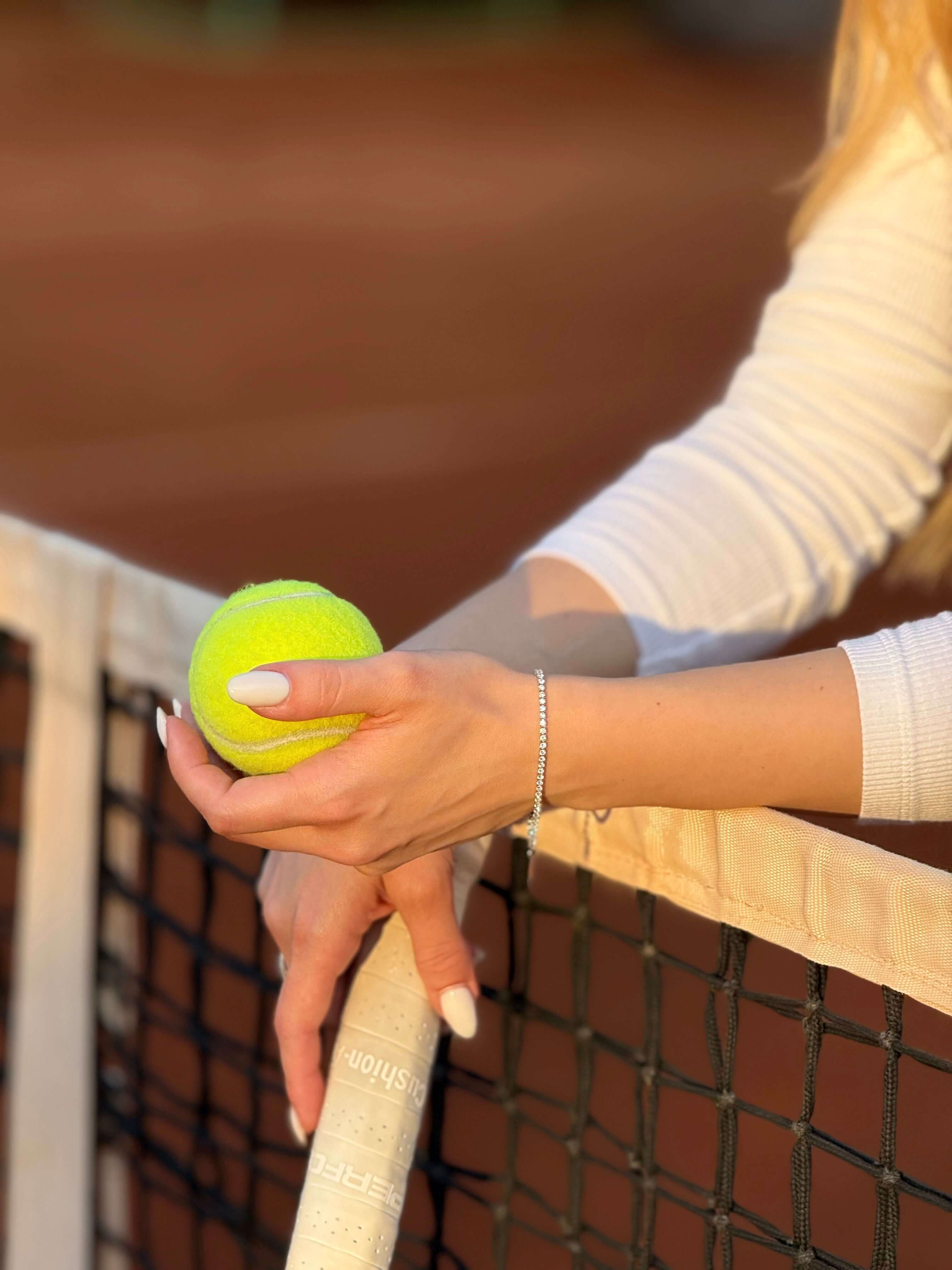 Жена държи тенис топка в ръка, докато се опира на тенис мрежа.