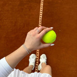 Ръка с тенис топка на тенис корт, носеща стилна гривна на фона на оранжево покритие.