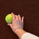 Ръка, държаща тенис топка на червена настилка, с елегантна тенис гривна.