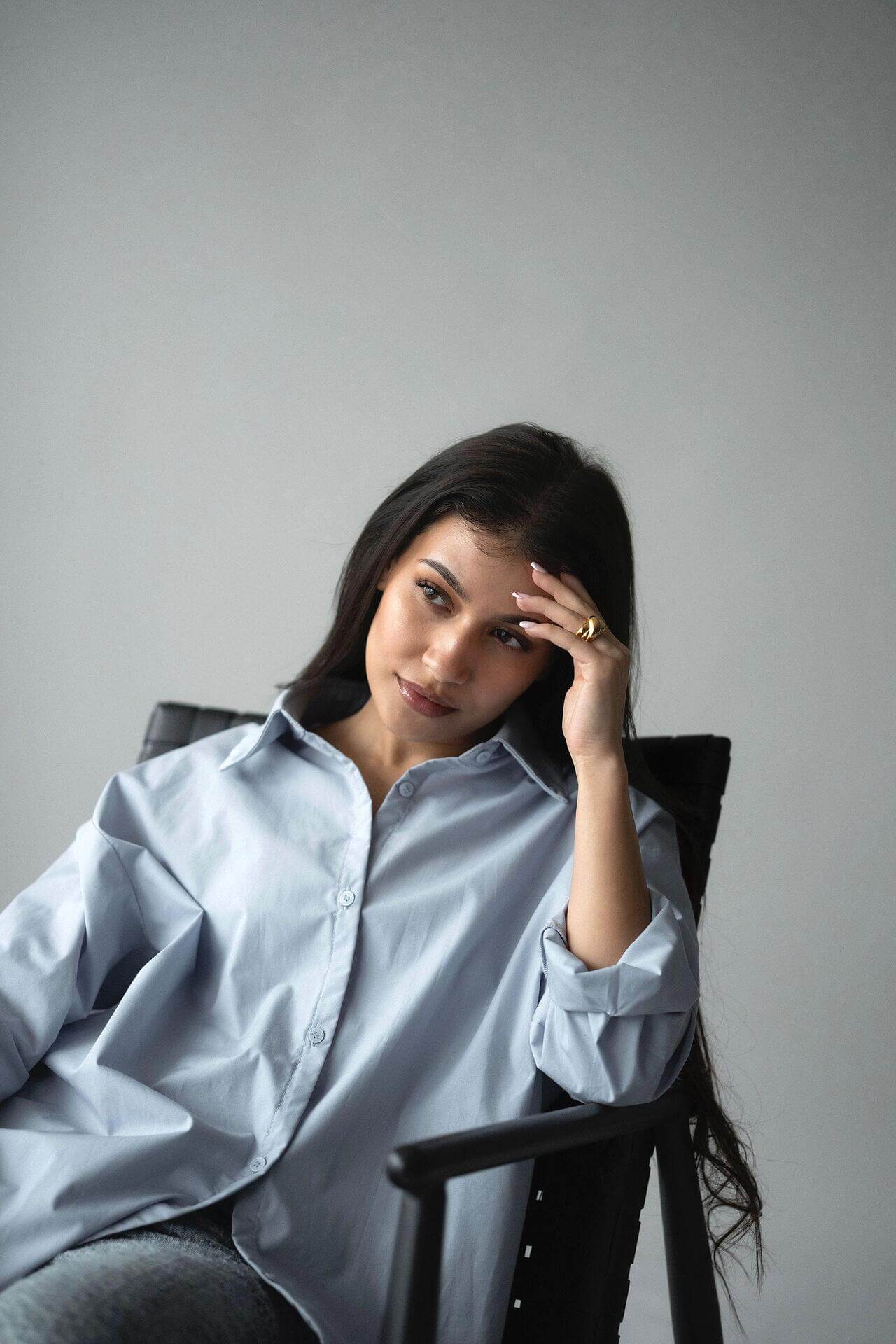 Woman in a blue shirt sitting on a chair, with long dark hair, appearing thoughtful.