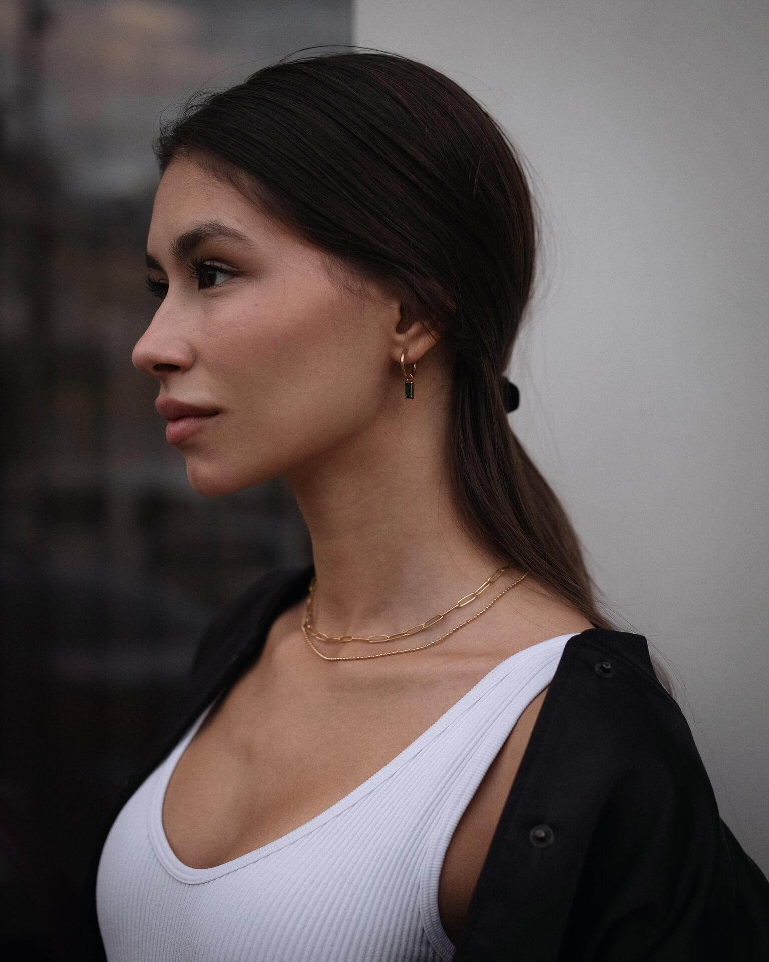 Млада жена с двойно златисто колие и обеци, подчертаваща елегантен стил на фона на неясен градски пейзаж.