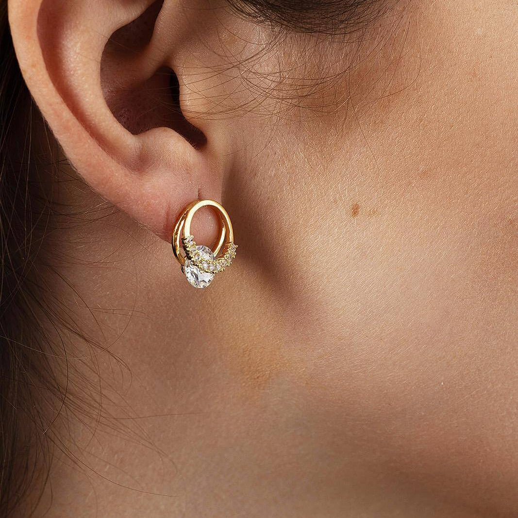 Обeци с блестящ камък в златист цвят, носени от жена, подчертаващи елегантността и стилa.