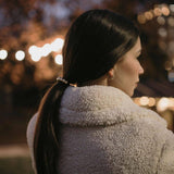 Жена с дълга коса, вързана в опашка, облечена в уютно палто на фона на вечерни светлини.