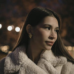 Млада жена с обеци "Криле" с 18К златно покритие, облечена в пухкаво палто, на фона на вечерна светлина.