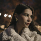 Млада жена с обеци "Криле" с 18К златно покритие, облечена в пухкаво палто, на фона на вечерна светлина.