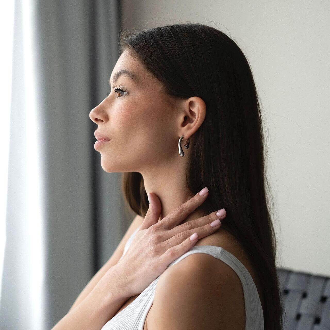 Elegant woman showcasing Обеци Течна Капка in gold, highlighting their shine and design by a window.