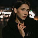 Модерна жена с обеци V-Shape, облечена в черно палто на фона на нощен град с размазани светлини.