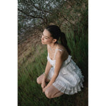 Млада жена с дълга коса седнала в зелена трева, носеща бяла рокля с ръкави, в естествена среда.