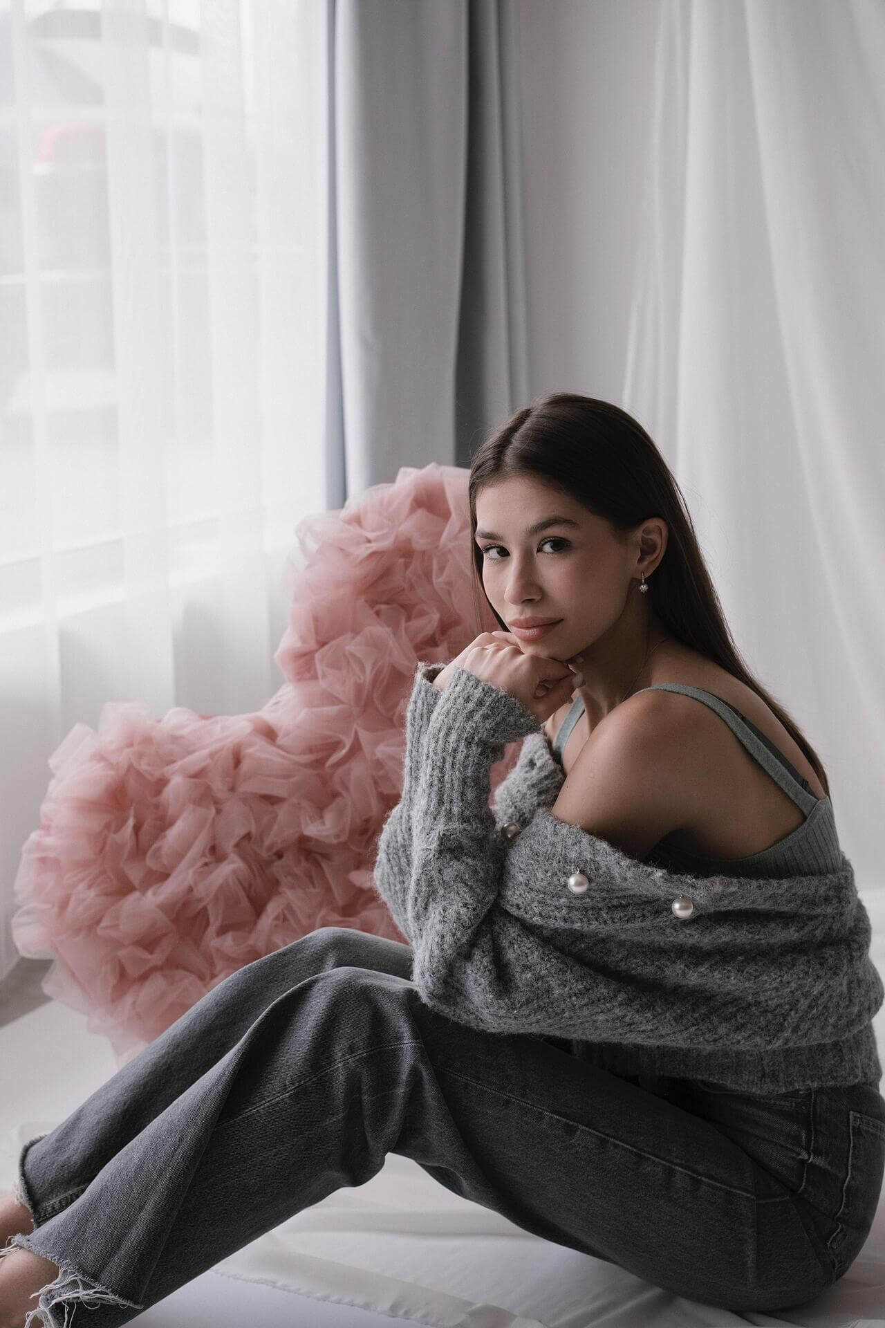 Млада жена сива блуза и дънки, усмихната на легло с розов сърцевиден възглавница.