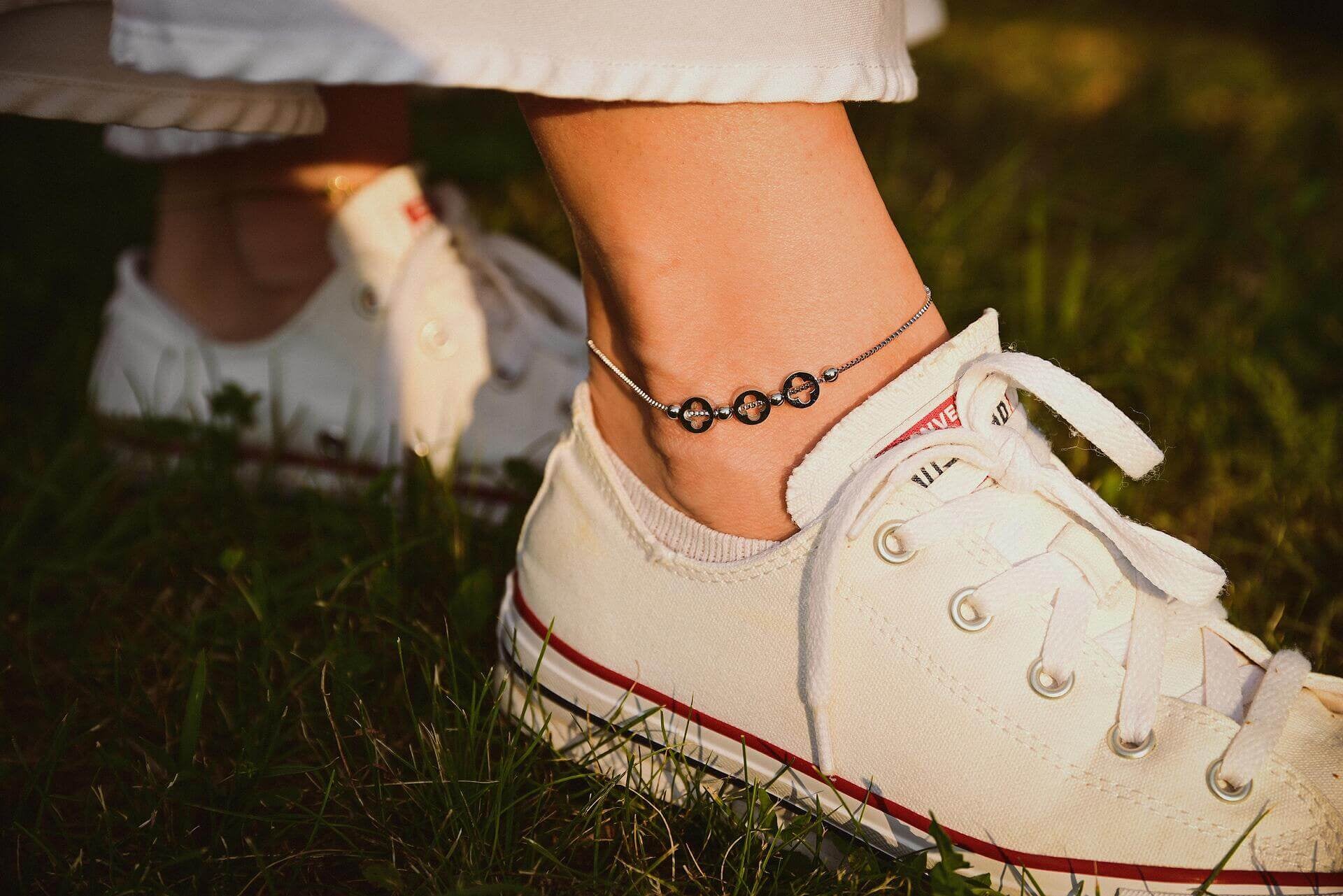 Модерна гривна за глезен от медицинска стомана с черни перли, носена с бели маратонки на зелена трева.