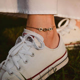 Гривна за глезен от медицинска стомана в златист цвят, носена с бели маратонки на фон от зелена трева.