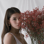 Млада жена с тъмна коса държи букет от червени цветя, носеща златни обеци и облечена в бяла тениска.