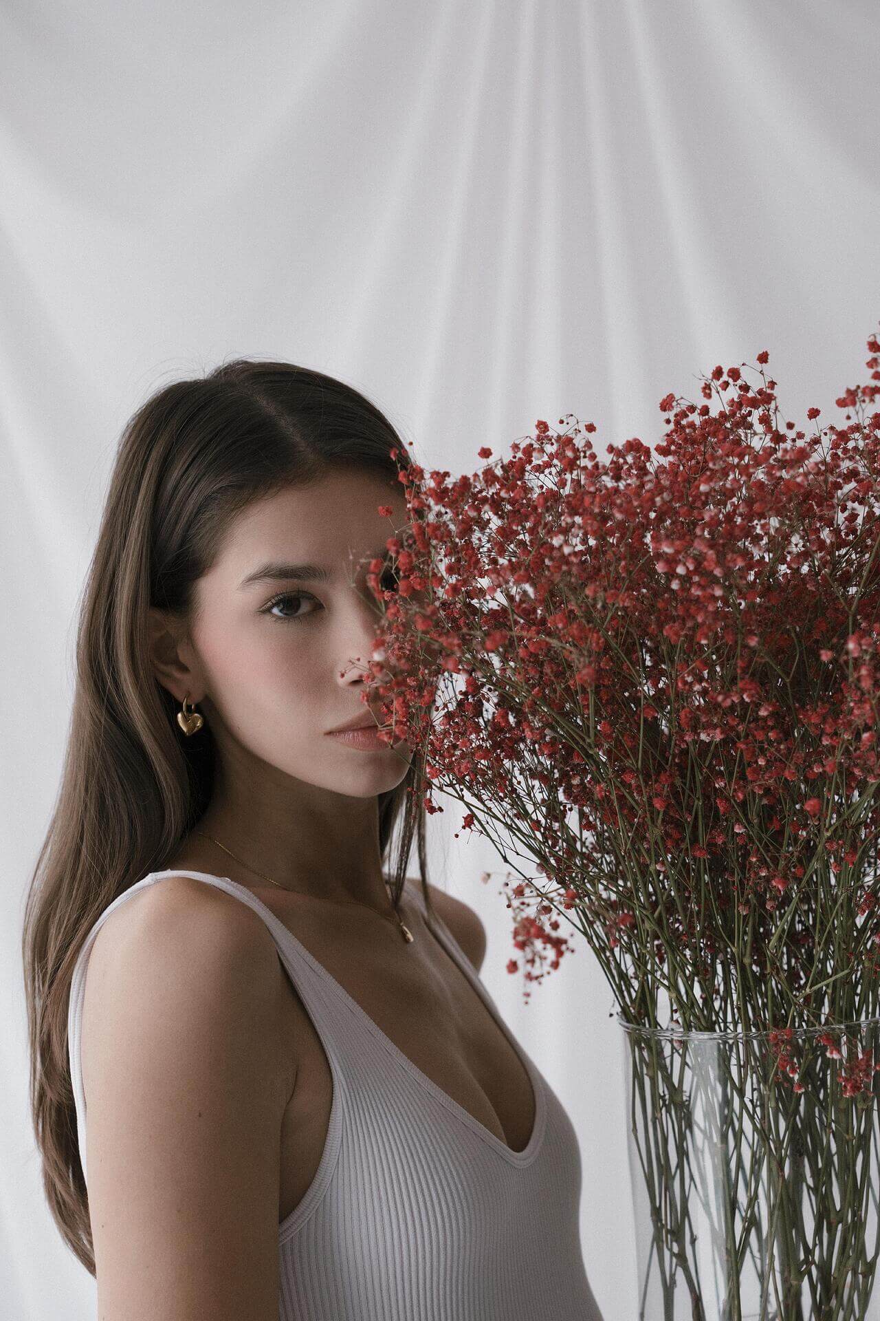 Млада жена с тъмна коса държи букет от червени цветя, носеща златни обеци и облечена в бяла тениска.