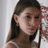 Жена с обеци сърце от 18K златно покритие, зад която се виждат червени цветя.