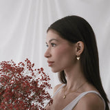 Жена с дълга тъмна коса, носеща обеци "Любов" с 18К златно покритие, държи червени цветя на фона на кремав текстил.