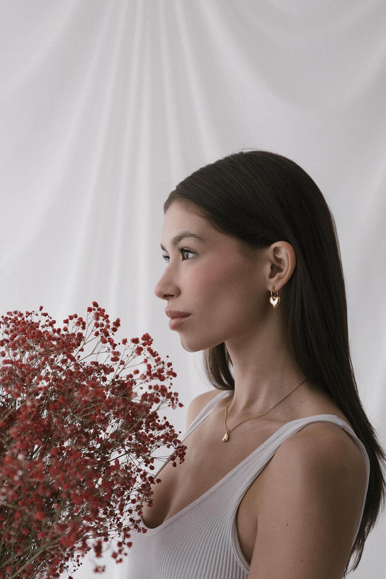 Жена с дълга тъмна коса, носеща обеци "Любов" с 18К златно покритие, държи червени цветя на фона на кремав текстил.