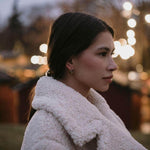 Млада жена в бяла палто с елегантни обеци, на фона на вечерни светлини.