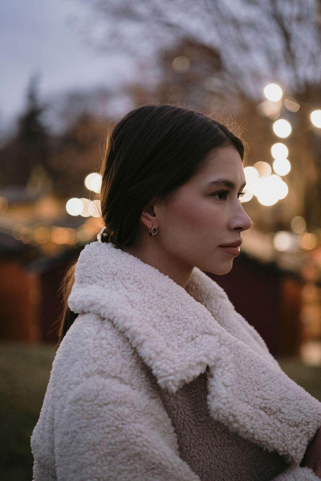 Млада жена в бяла палто с елегантни обеци, на фона на вечерни светлини.