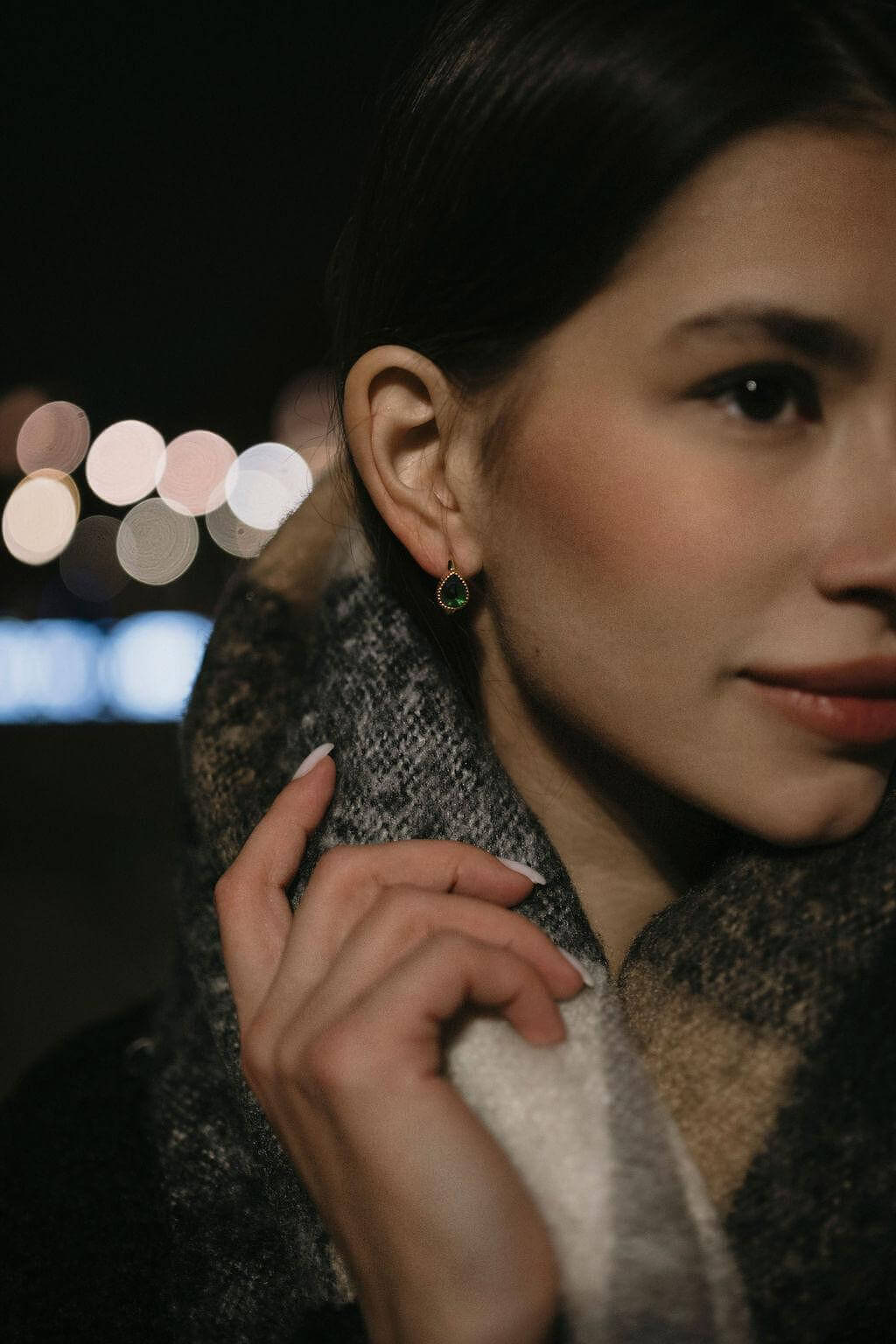 Млада жена с златисти обеци с многонюансни кристали, облечена в тъмен шал, на фона на размазани светлини.