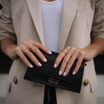 Жена с изискано облекло държи черен портфейл, носейки златисти пръстени и нежно маникюр.