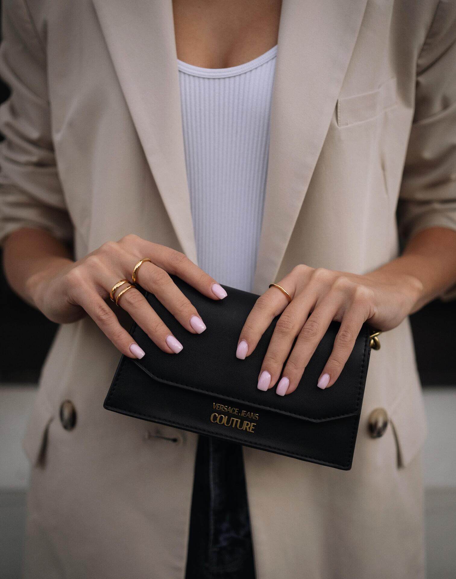 Жена с изискано облекло държи черен портфейл, носейки златисти пръстени и нежно маникюр.