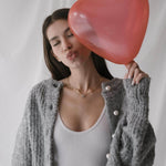 Млада жена в серо-сив пуловер с перли, държи червена сърцеформа балон.