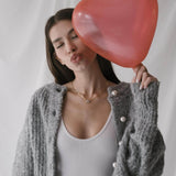 Млада жена в серо-сив пуловер с перли, държи червена сърцеформа балон.