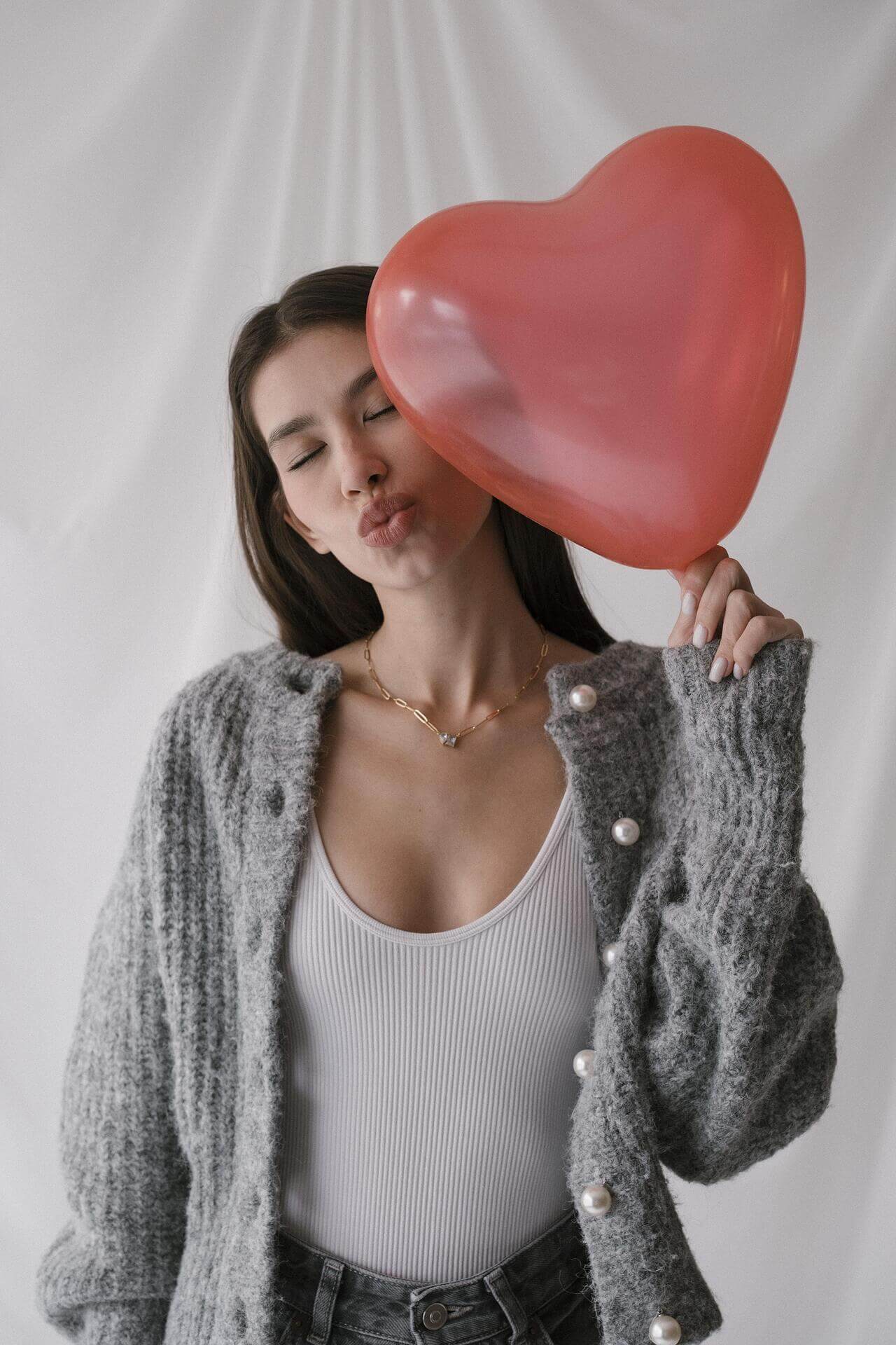 Млада жена в серо-сив пуловер с перли, държи червена сърцеформа балон.